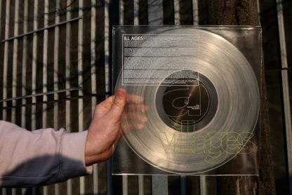 Picture of a hand, holding the second press of the Villages "ILL AGES" LP in front of an iron gate